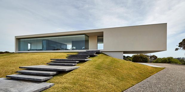 Low-slung minimalist contemporary house set into a gently sloping landscaped site. The elongated flat-roofed form has a clean pale rendered facade, extensive floor-to-ceiling glazing, and a dramatic open-riser dark stone or concrete stair rising across the lawn to the entrance. Gravel paving and sparse coastal vegetation surround the residence.