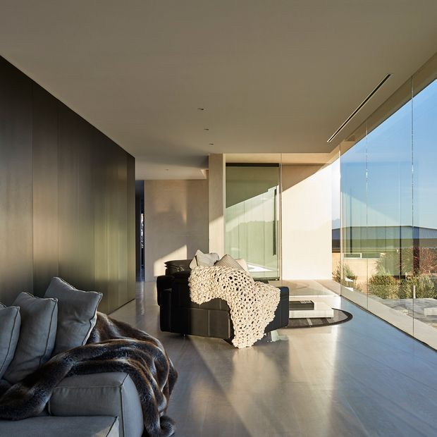 A refined contemporary living room with floor-to-ceiling glazing along one side, opening to a bright exterior outlook. The room features a low modular dark sofa in the foreground, a central dark lounge chair with a cream knitted throw, smooth light-toned flooring, a dark timber or wood-look feature wall, recessed ceiling lighting, and strong natural light. The space has a long, open linear composition and minimal built-in detailing.