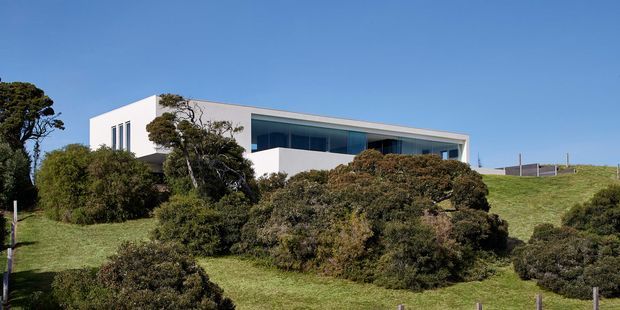 Exterior view of a modern, low-profile white house set within a gently sloping, landscaped coastal-looking site. The building has crisp geometric massing, a flat roof, extensive dark-framed horizontal glazing, and a large recessed upper-level terrace or balcony. Dense native-looking shrubs and mature trees partially screen the lower facade.