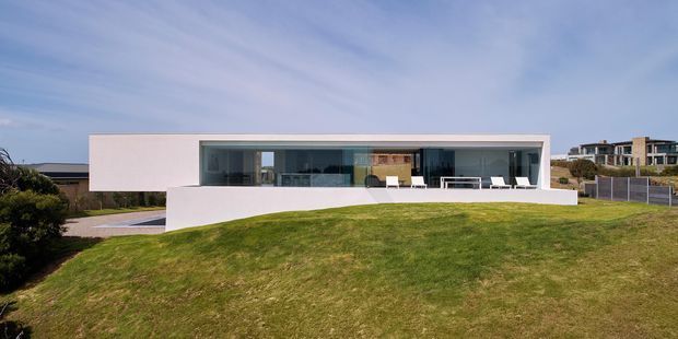 A low, minimalist modern residence viewed from the landscaped front lawn. The building has a long rectangular form, crisp white rendered walls, a broad flat roof, extensive dark-framed floor-to-ceiling glazing, and a projecting white terrace or plinth. Neighbouring contemporary homes are visible in the background.