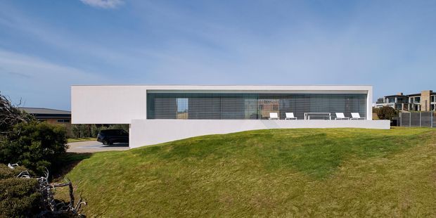 A wide exterior view of a minimalist, single-storey white modern house set into a gently sloping grass-covered site. The long rectangular facade features extensive floor-to-ceiling glazing with horizontal screening, a projecting white roof or wall form, and a sheltered carport opening at the left. A low white boundary or retaining wall runs across the foreground, with neighbouring buildings visible in the distance.