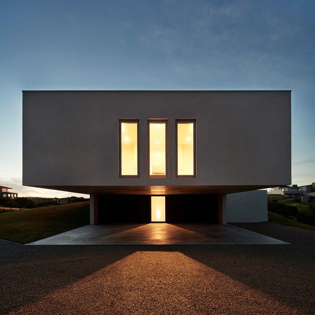 Minimalist contemporary two-storey rectangular house photographed at dusk. The dark, smooth, box-like upper volume projects over an open ground-level carport, with three narrow illuminated vertical windows and a single glowing entry door. A broad concrete driveway and sparse grassy suburban surroundings reinforce the stark, geometric composition.