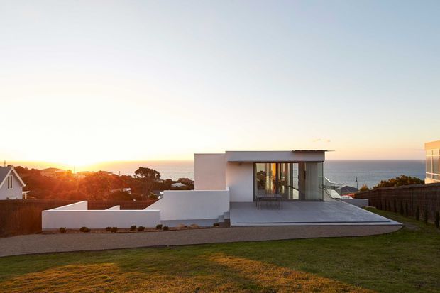 Exterior view of a contemporary flat-roofed residence at sunset, positioned on a gently sloping site with expansive ocean views. The house has crisp white rendered walls, broad dark-framed glazing, a projecting upper roof slab, and a large paved terrace. A lawn and gravel foreground lead toward neighbouring homes and the coastline.