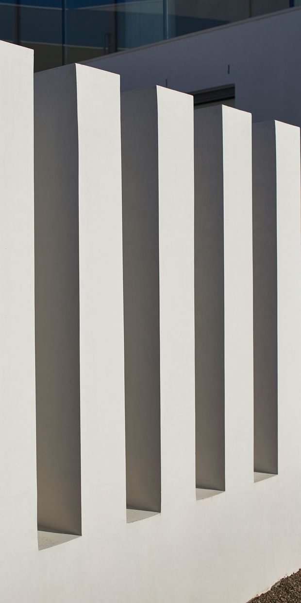 Close-up architectural exterior detail showing a row of tall, evenly spaced white vertical fins or projecting walls casting deep shadows, with dark glazed building surfaces and a pale rendered wall behind. The composition is geometric and minimalist, emphasizing repetition, light, and shadow.