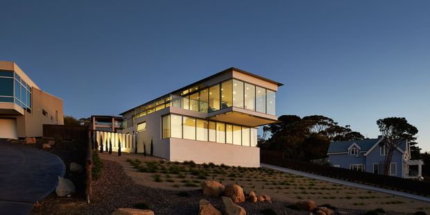 Twilight exterior view of a contemporary multi-level house on a sloping landscaped site. The building features a flat roof, strong rectilinear volumes, extensive floor-to-ceiling glazing, projecting upper-level forms, and pale rendered walls with dark framing. A neighbouring coastal-style house and mature trees are visible in the background.
