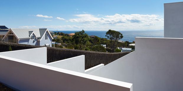 Exterior architectural view from a modern white rendered building, with stepped parapet walls and clean geometric forms in the foreground. Beyond are residential rooftops, coastal vegetation, and a clear ocean horizon under a bright blue sky.