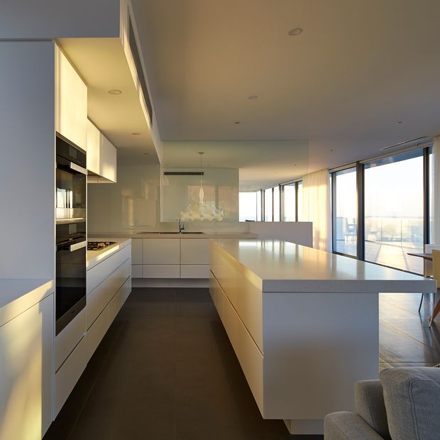 A sleek open-plan kitchen with white flat-panel cabinetry, integrated black appliances, a broad central island, and a pale stone benchtop. The kitchen opens toward a bright living area with full-height glazing and expansive views. Recessed ceiling lighting, dark flooring, and minimal detailing create a refined contemporary interior.