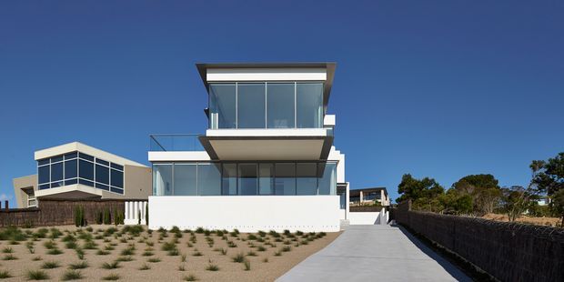 A contemporary multi-level coastal residence viewed from the street, with crisp white walls, extensive floor-to-ceiling glazing, projecting box-like volumes, and a flat roof. A broad concrete driveway leads toward the house, while a sparsely planted front landscape and dark boundary fencing frame the property beneath a clear blue sky.