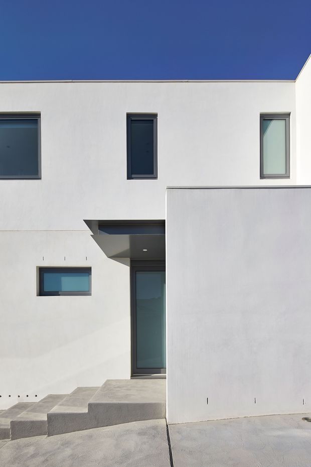A minimalist modern residential facade with crisp white rendered walls, dark-framed windows and a tall glazed entry door. The asymmetrical, cubic composition includes a projecting upper volume, a recessed entrance canopy, broad concrete steps and a plain foreground wall. The image is photographed outdoors in strong daylight against a clear blue sky.