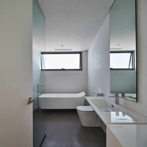 A bright, contemporary bathroom viewed through an open door, featuring a freestanding white bathtub, wall-mounted toilet, long white vanity with integrated basin and chrome tapware, a large mirror, frameless glass shower enclosure, dark floor tiles, pale textured walls, and a high horizontal awning window.