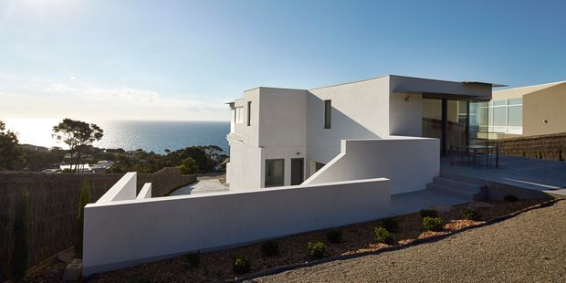 Exterior view of a contemporary multi-level white-rendered home on a coastal hillside, with strong geometric forms, flat rooflines, terraces, broad glazing, and low white boundary walls. The ocean and coastal landscape are visible beyond the property.