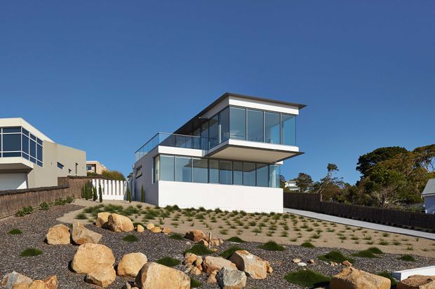 Exterior view of a contemporary multi-level house on a landscaped sloping site. The building has crisp white rendered walls, extensive floor-to-ceiling glazing, projecting upper-floor volumes, a flat roof, and glass balustrades. The foreground features a low-maintenance gravel and rock landscape with sparse planted greenery, while neighbouring homes and mature trees are visible around the property.