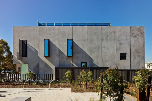 Exterior view of a stark, rectilinear modern building with a monolithic grey concrete facade, narrow blue-tinted vertical windows, deep shadow lines, dark vertical fencing, and a flat roof with a raised glazed roof element. The foreground includes landscaped planting and bicycle racks.