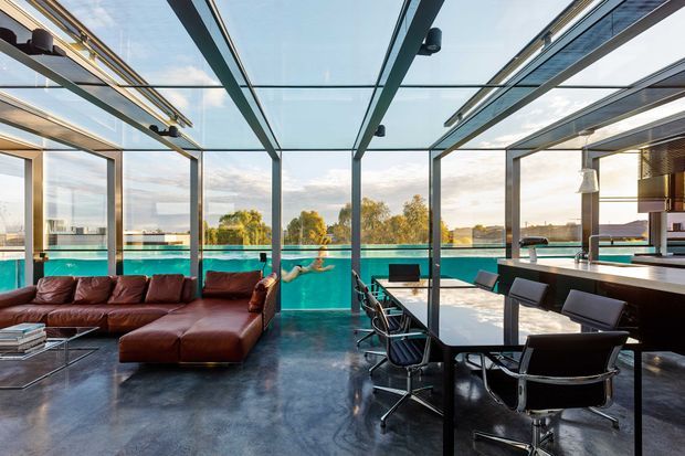A spacious, open-plan contemporary living and dining area with floor-to-ceiling glazing and a transparent glazed roof structure. The room contains a dark leather sectional sofa, a polished dark floor, a large dining table with black leather office-style chairs, and a kitchen or bar counter at right. Turquoise pool water and landscaped surroundings are visible beyond the glazing, creating a bright indoor-outdoor connection.