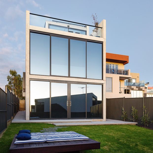 Rear exterior view of a compact contemporary multi-storey residence with a strongly framed glazed facade, full-height dark-framed windows and doors, rooftop terrace or balcony, and a small landscaped lawn with an outdoor daybed in the foreground. Adjacent modern homes are visible in the background.