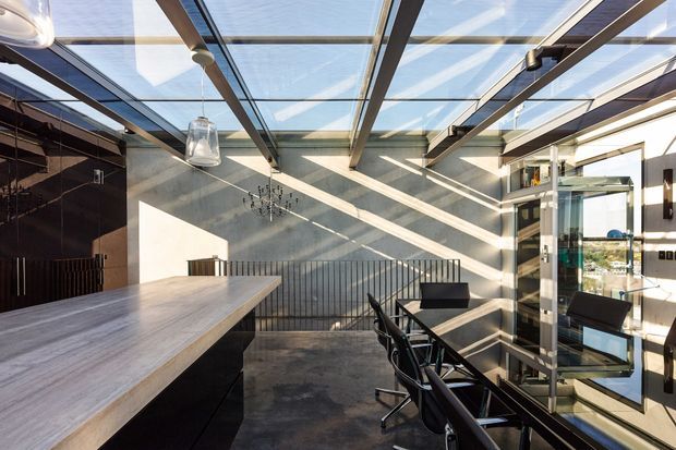 A contemporary open-plan kitchen and dining area beneath a dramatic glazed roof. The space features a large pale timber island with dark cabinetry, a long dining table with black chairs, polished concrete flooring, dark reflective cabinetry, exposed dark structural beams, pendant lighting, and extensive glazing that fills the room with sunlight. A glazed door and shelving connect visually to the exterior.