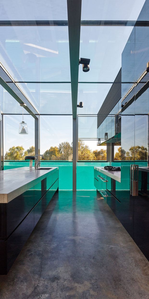 A contemporary kitchen beneath a transparent glazed roof, with strong exposed structural framing and extensive glazing overlooking trees and a turquoise pool. The kitchen features dark teal/black glossy fixed cabinetry, pale stone benchtops, a central island with integrated sink and tap, a separate cooktop run, pendant lighting, and a dark polished concrete floor. Reflections in the glazing show the surrounding structure and cabinetry.