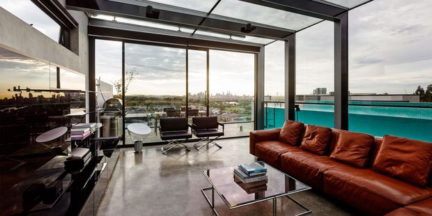 A spacious contemporary living room with floor-to-ceiling steel-framed glazing and a glazed roof, opening to a broad city and water view. The room features polished concrete flooring, a large cognac-brown leather sectional sofa, black lounge chairs, a glass coffee table, and dark built-in cabinetry or media joinery along the left wall. A turquoise swimming pool or terrace edge is visible outside.