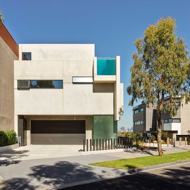Exterior street view of a modern multi-level residence with a minimalist cubic form, smooth pale rendered walls, narrow horizontal windows, a recessed double garage, and a turquoise glazed balcony or feature opening. The property sits along a landscaped suburban street with neighbouring contemporary homes and a visible urban skyline in the distance.