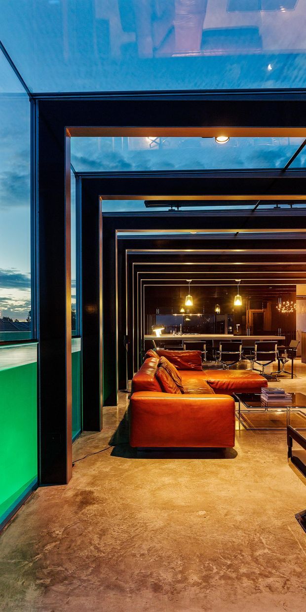 A dramatic interior view through a sequence of black-framed glazed openings into an open-plan living, dining and kitchen space. The room features a large cognac leather sectional sofa, polished concrete flooring, dark walls and structural framing, a long kitchen island with pendant lights, a dining table, and warm layered lighting. Extensive glazing and repeated rectangular frames create a strong tunnel-like perspective, with dusk visible outside.