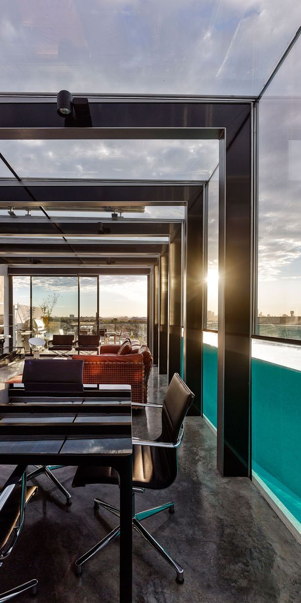 A dramatic contemporary interior with a long perspective of dark metal-framed glazed bays and repeated skylight-like roof sections. The space combines a black dining or work table with leather office-style chairs, a lounge seating area, polished dark flooring, and expansive glazing overlooking a city skyline. A vivid turquoise pool or water feature runs alongside the glazed wall, while warm low sunlight creates strong reflections and contrast.