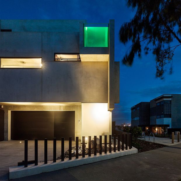A dusk exterior photograph of a contemporary multi-level urban residence. The building has a minimalist cubic form with smooth concrete-rendered walls, projecting volumes, narrow horizontal openings, a glowing green upper-level window, and a dark recessed garage. Black bollards line the driveway and pedestrian edge, while neighbouring modern buildings are visible in the background.
