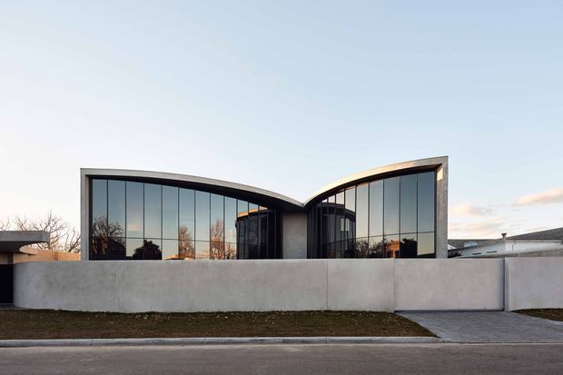 Exterior street view of a sculptural contemporary residence with two sweeping curved roof forms meeting at a central valley. The facade is dominated by tall floor-to-ceiling dark-framed glazing, with smooth pale rendered boundary walls, a paved driveway, and restrained landscaping. The low, symmetrical composition has a clean modern character and reflects the surrounding trees in the glass.
