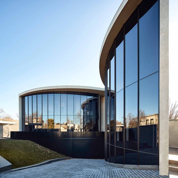 Exterior architectural photograph of a contemporary building composed of sweeping curved forms, extensive floor-to-ceiling blue-tinted glazing, slender dark window frames, and pale concrete or rendered structural elements. A paved forecourt, dark privacy fencing, and landscaped grass surround the facade under a clear sky.