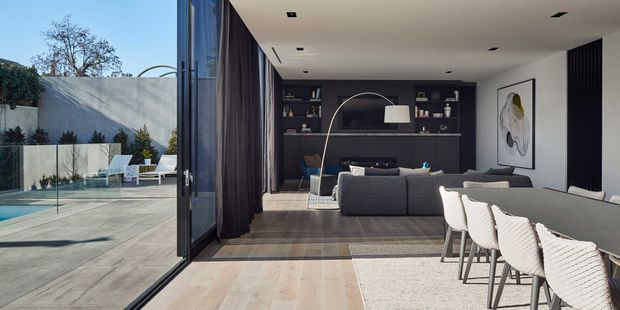 Contemporary open-plan living and dining space opening onto a paved courtyard through expansive floor-to-ceiling glazing. The room features a dark built-in media and display wall, charcoal sectional sofa, sculptural floor lamp, pale upholstered dining chairs, wide timber flooring, a large light rug, recessed ceiling lights, and dark full-height curtains. A swimming pool and outdoor seating are visible beyond the glazing.