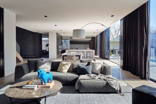 Open-plan contemporary living area with a large grey sectional sofa and ottoman in the foreground, a round marble coffee table, and floor-to-ceiling glazing dressed with dark curtains. A kitchen with a central island and pendant light is visible behind the living space, along with a dark textured feature wall and stair opening. The room is bright and refined, with strong indoor-outdoor visual connection through the glazing.