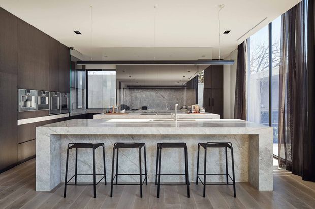 A spacious contemporary kitchen featuring a large stone island with four black metal bar stools, dark timber cabinetry, integrated appliances, and a stone backsplash. Floor-to-ceiling glazing and dark curtains bring in abundant natural light, while recessed and pendant lighting create a clean, refined interior.