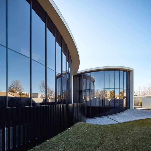 Exterior view of a contemporary curved building with extensive floor-to-ceiling glazing, dark vertical metal screening, and a pale curved roof edge. The structure forms rounded wings around a paved entry or courtyard area, with lawn in the foreground and suburban surroundings visible through the glass.