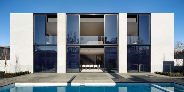 Front-on exterior view of a minimalist two-storey modern house with a symmetrical white rendered facade, tall dark-framed floor-to-ceiling glazing, and a central recessed entry. A swimming pool and paved terrace occupy the foreground, while the interior is visible through the glazed opening.