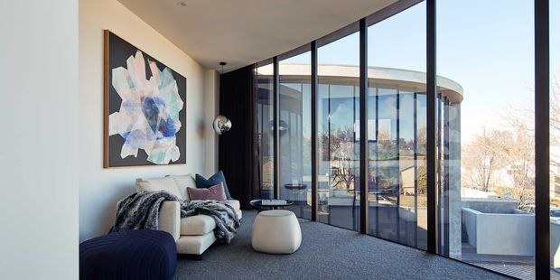 A refined contemporary living room with floor-to-ceiling black-framed glazing wrapping around a curved exterior form. The space contains a cream lounge chair, dark upholstered seating, a small light ottoman, a sculptural floor lamp, and abstract wall art, with a dark carpeted floor and strong views to the surrounding landscape and adjacent curved structure.