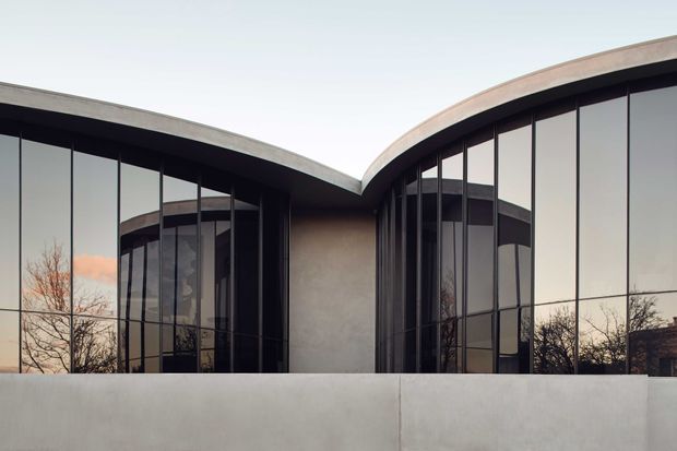 Exterior architectural photograph of a sculptural modern building composed of two curved, wing-like volumes with sweeping concrete roof edges, dark slender window frames, extensive reflective glazing, and a smooth pale concrete privacy wall in the foreground. The image has a restrained monochromatic palette and emphasizes the building’s curved geometry and facade composition.