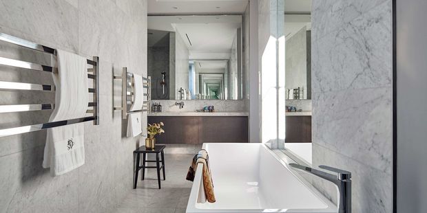 Contemporary bathroom featuring a freestanding white bathtub, large marble-look wall surfaces, extensive mirrored cabinetry, a floating dark wood vanity with double basins, and sleek chrome fixtures. A ladder-style heated towel rail and stool add functional detail. The long sightline and repeated reflections create a bright, refined, high-end atmosphere.