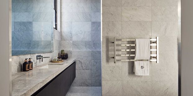 Contemporary bathroom interior featuring a long stone vanity with dark built-in cabinetry, wall-mounted tapware, pale marble-look wall and floor tiles, and a wall-mounted heated towel rail with folded towels. A narrow vertical window brings in natural light, while small toiletries sit on the vanity.