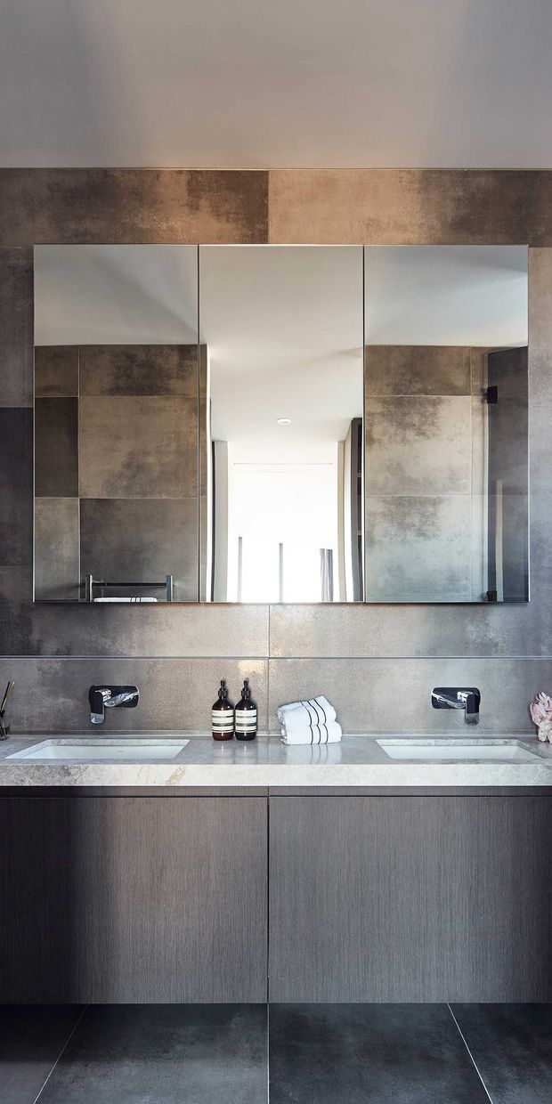 Contemporary double-vanity bathroom with a wall-mounted dark wood-grain vanity, two undermount basins, chrome wall-mounted taps, large three-panel mirrored cabinet, and large-format mottled grey-brown wall and floor tiles. The space has a restrained, refined palette with integrated lighting and clean linear detailing.
