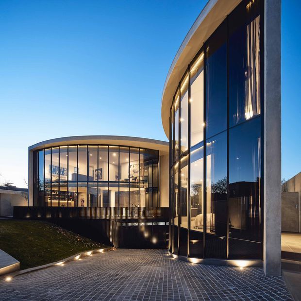 A contemporary multi-level building photographed at dusk, with sweeping curved forms, extensive floor-to-ceiling glazing, dark vertical fins, and exposed concrete columns. A paved forecourt leads toward the illuminated interior, where a large open-plan space and staircase are visible through the glass. The design emphasizes sculptural geometry, transparency, and integrated architectural lighting.