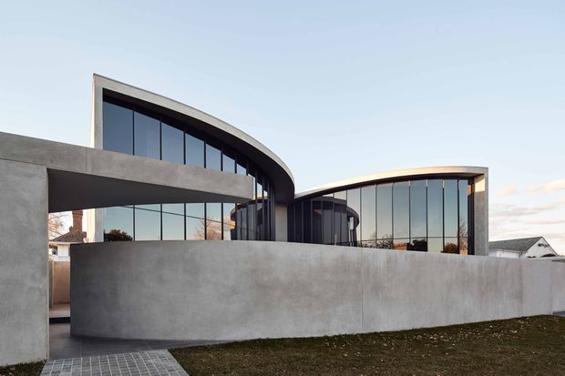Exterior view of a contemporary multi-level building composed of curved concrete forms and sweeping rooflines, with extensive dark-framed floor-to-ceiling glazing. A tall solid concrete boundary wall screens the lower level, while a paved walkway and lawn occupy the foreground. The design has a sculptural, restrained architectural character.