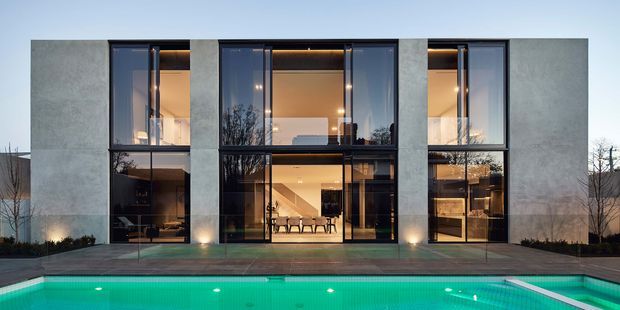 Front exterior view of a contemporary multi-storey residence with a rectilinear concrete facade and extensive dark-framed floor-to-ceiling glazing. Warm interior lighting reveals living and dining areas, while a timber-toned terrace and illuminated swimming pool occupy the foreground.