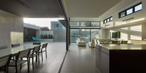 Spacious contemporary open-plan interior with a dining area, kitchen island and living room opening onto a large paved courtyard and swimming pool. Floor-to-ceiling glazing, clerestory windows, and extensive horizontal louvres create strong indoor-outdoor connection and filtered sunlight. The space uses large pale grey floor tiles, dark cabinetry, white walls, and a slatted ceiling feature.