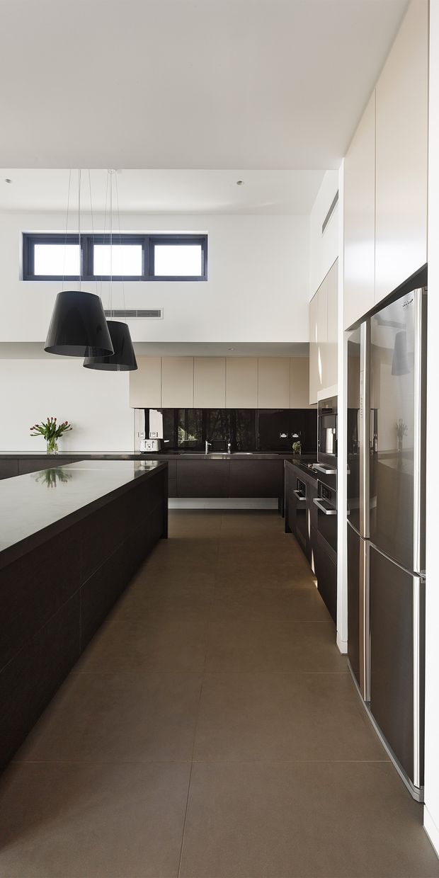 A sleek contemporary kitchen viewed along a long central aisle, featuring a large dark island with a pale stone benchtop, dark lower cabinetry, pale upper cabinetry, a glossy black splashback, integrated stainless-steel appliances, and large dark pendant lights. White walls and ceiling, high clerestory-style windows, and warm large-format floor tiles create a bright, refined space.