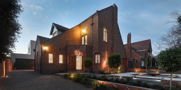 Exterior view of a substantial red-brick residence with strong ecclesiastical and Gothic Revival influences. The building features steep gabled forms, tall arched windows, a prominent brick chimney, dark tiled roofs, timber or dark-framed doors and windows, and carefully landscaped front grounds with a paved driveway and low brick-edged garden beds. Warm exterior lighting highlights the facade at dusk.