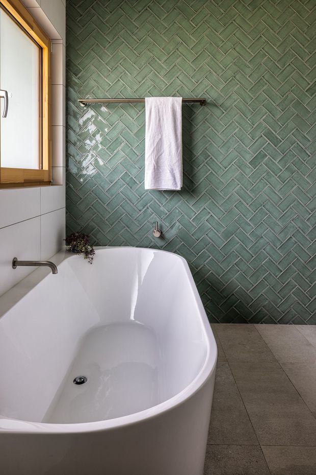 A contemporary bathroom featuring a freestanding white bathtub, wall-mounted chrome tapware, a timber-framed frosted window, and a full-height glossy green herringbone tile feature wall. Large grey-beige floor tiles and a simple dark metal towel rail complete the calm, refined interior.