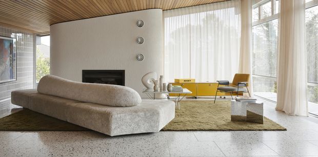 A bright mid-century-inspired living room with a curved textured sofa, low rectangular coffee table, yellow sideboard, sculptural lounge chair, and large sheer-curtained windows. The room features a warm timber slatted ceiling, white plaster walls with a linear fireplace and circular wall details, and a terrazzo or concrete-look floor beneath a muted area rug.