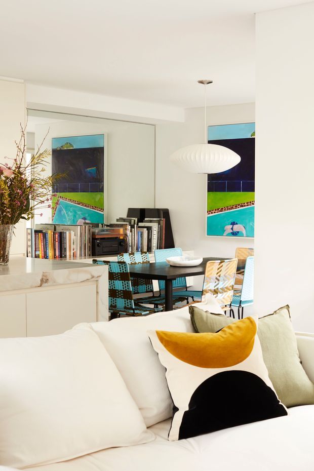 Bright open-plan living and dining area with a white sofa in the foreground, colourful geometric cushions, a dark dining table with patterned upholstered chairs, large vibrant abstract artwork, a sculptural white pendant light, mirrored cabinetry or wall panels, and a light stone kitchen island edge.