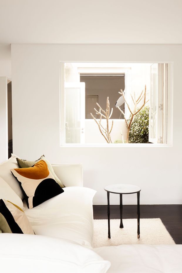 Bright, minimalist interior corner with a white upholstered sofa or daybed, graphic black-and-orange cushions, a small round black side table, and a light rug over dark timber flooring. White walls and a large open internal window or opening provide strong natural light, with a planted courtyard visible beyond.