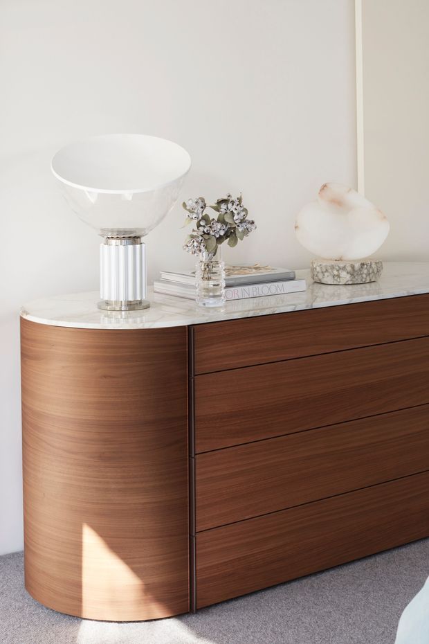 A refined interior vignette showing a curved mid-century-style dark timber sideboard with integrated drawers, topped by a pale stone or marble surface. A sculptural white table lamp, glass vase with flowers, books, and a rounded decorative object create a softly styled composition against an off-white wall. Grey carpet and warm natural light contribute to an elegant, calm atmosphere.