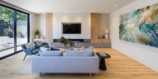 Bright contemporary living room with a large light-grey sectional sofa, blue accent chairs, timber flooring, a wall-mounted television, built-in timber joinery, a textured stone fireplace surround, and expansive black-framed glazing opening toward a leafy outdoor area. A large abstract artwork provides a colourful feature on the right wall.
